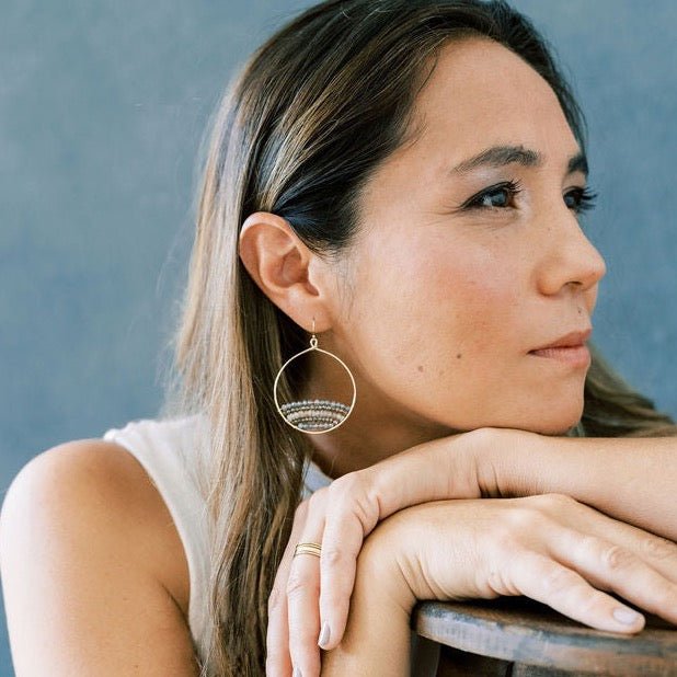 Woman with long dark hair wearing a white tank with gold and gemstone statement 2 inch hoop earrings with rows of zircon, labradorite, and smokey topaz gemstones. Harper Earrings by Sarah Cornwell Jewelry