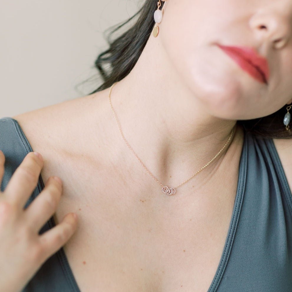 Woman's neckline with dark hair and red lipstick wearing gray tank top with gold necklace with 5 tiny gold rings and pearl and textured disc drop earrings. Honor Necklace by Sarah Cornwell Jewelry