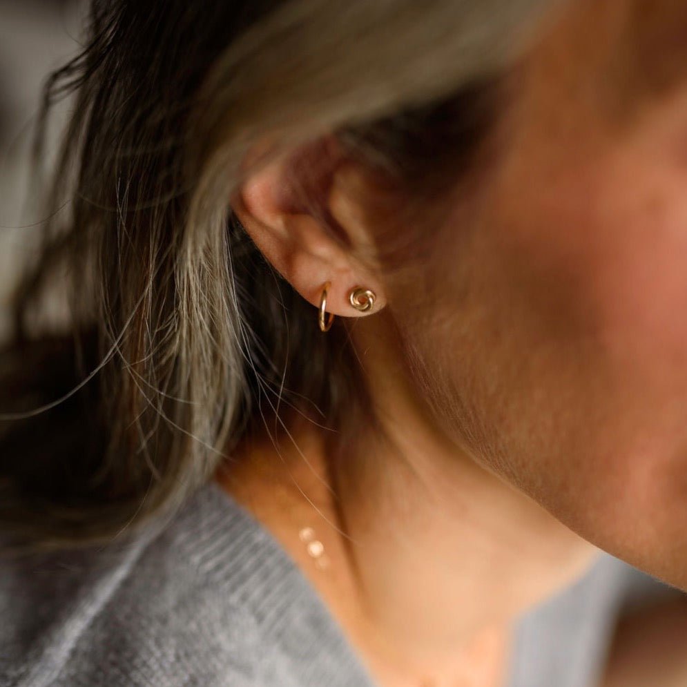 Woman with blonde hair wearing 6 mm gold love knot stud earrings and gold huggie hoop earrings. Love Knot Studs by Sarah Cornwell Jewelry