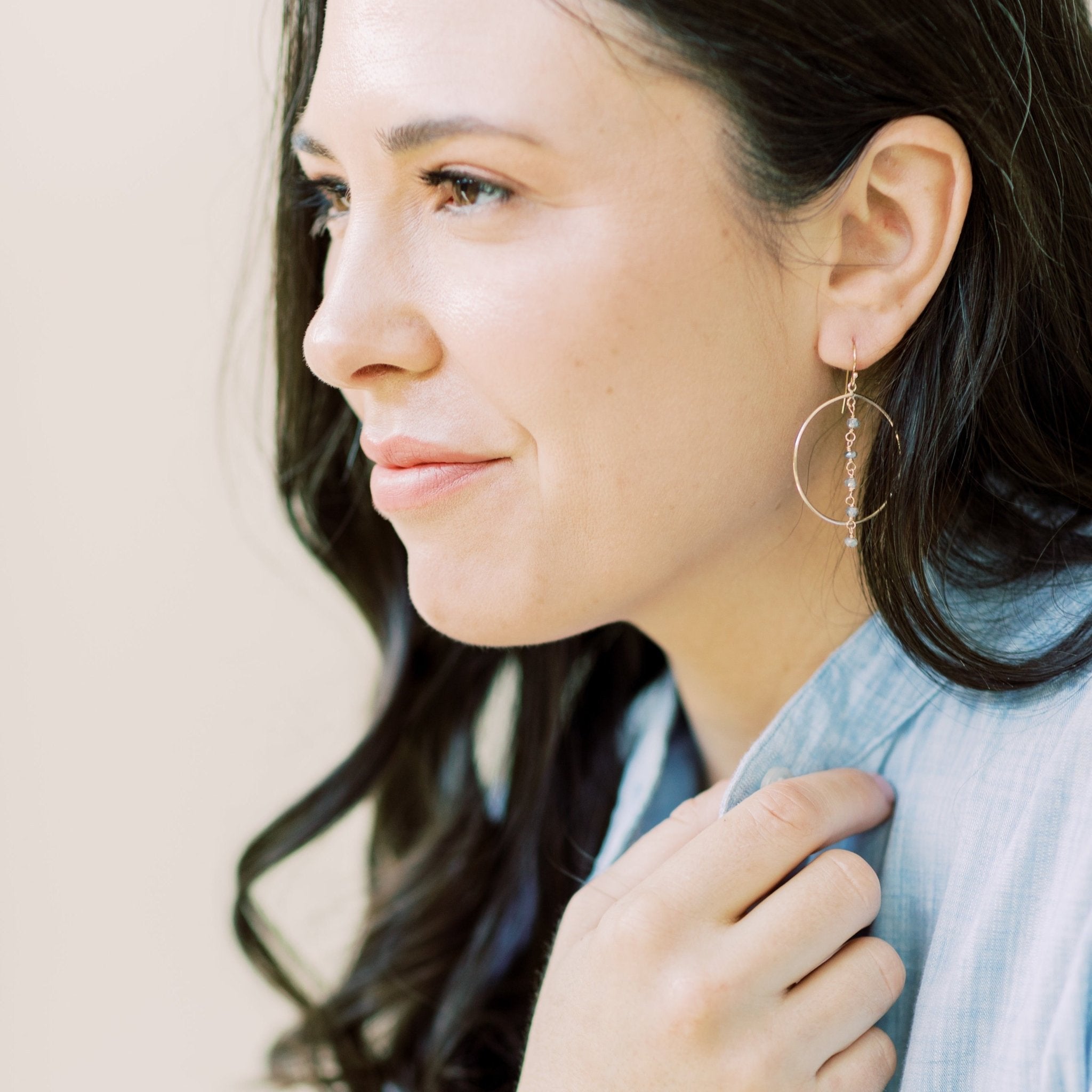 Gold Nantucket Earrings by Sarah Cornwell Jewelry. Dark haired woman wearing chambray shirt and sparkly statement gold hoop earrings with a strand of 5 wire wrapped labradorite gemstones down the middle.