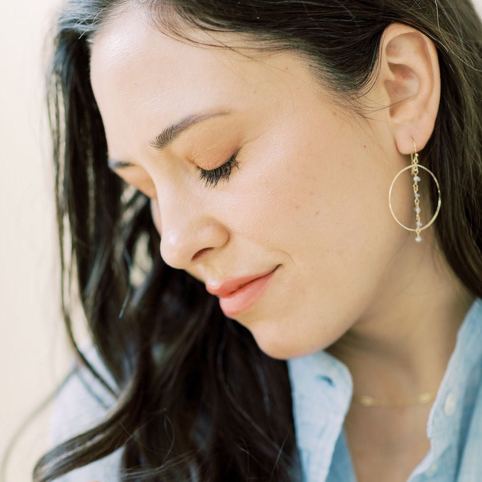 Gold Nantucket Earrings by Sarah Cornwell Jewelry. Side view of dark haired woman with eyes closed wearing chambray shirt and sparkly statement gold hoop earrings with a strand of 5 wire wrapped labradorite gemstones down the middle.