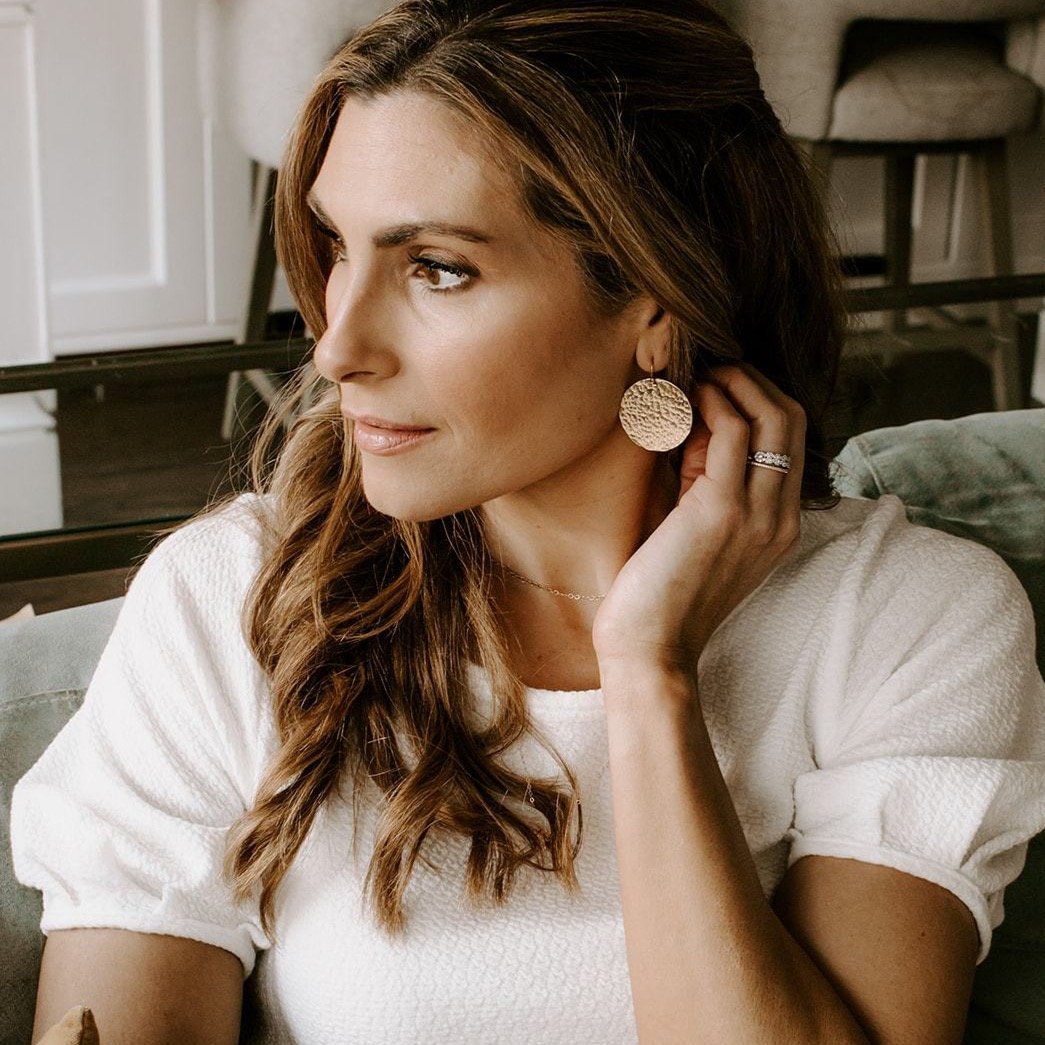 Gold Phoebe Earrings by Sarah Cornwell Jewelry. Blonde haired woman sitting on sofa wearing white short sleeved top and shimmery, textured 1.25 inch gold disc statement earrings and gold chain necklace .