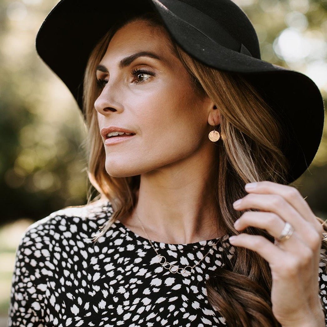 Gold Rachel Earrings by Sarah Cornwell Jewelry. Side view of blonde woman wearing a black hat and black and white top and shimmery, dainty gold 1/2 inch textured disc earrings and gold 5 circle necklace.