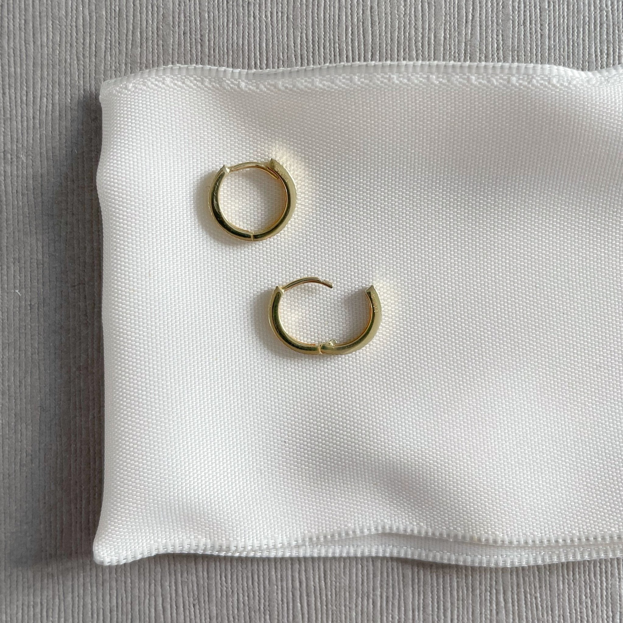 Solid Gold Ruth Huggies by Sarah Cornwell Jewelry. Solid gold tiny hoop huggie earrings with one open on a white and gray background.