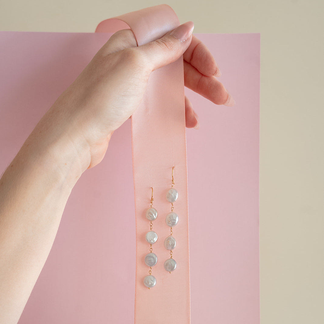 Hand holding pink ribbon with a pair of coin pearl statement earrings on a pink background