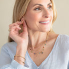 A woman with blonde hair wearing a light blue blouse and delicate gold jewelry, including layered necklaces and earrings, posing against a neutral beige background.