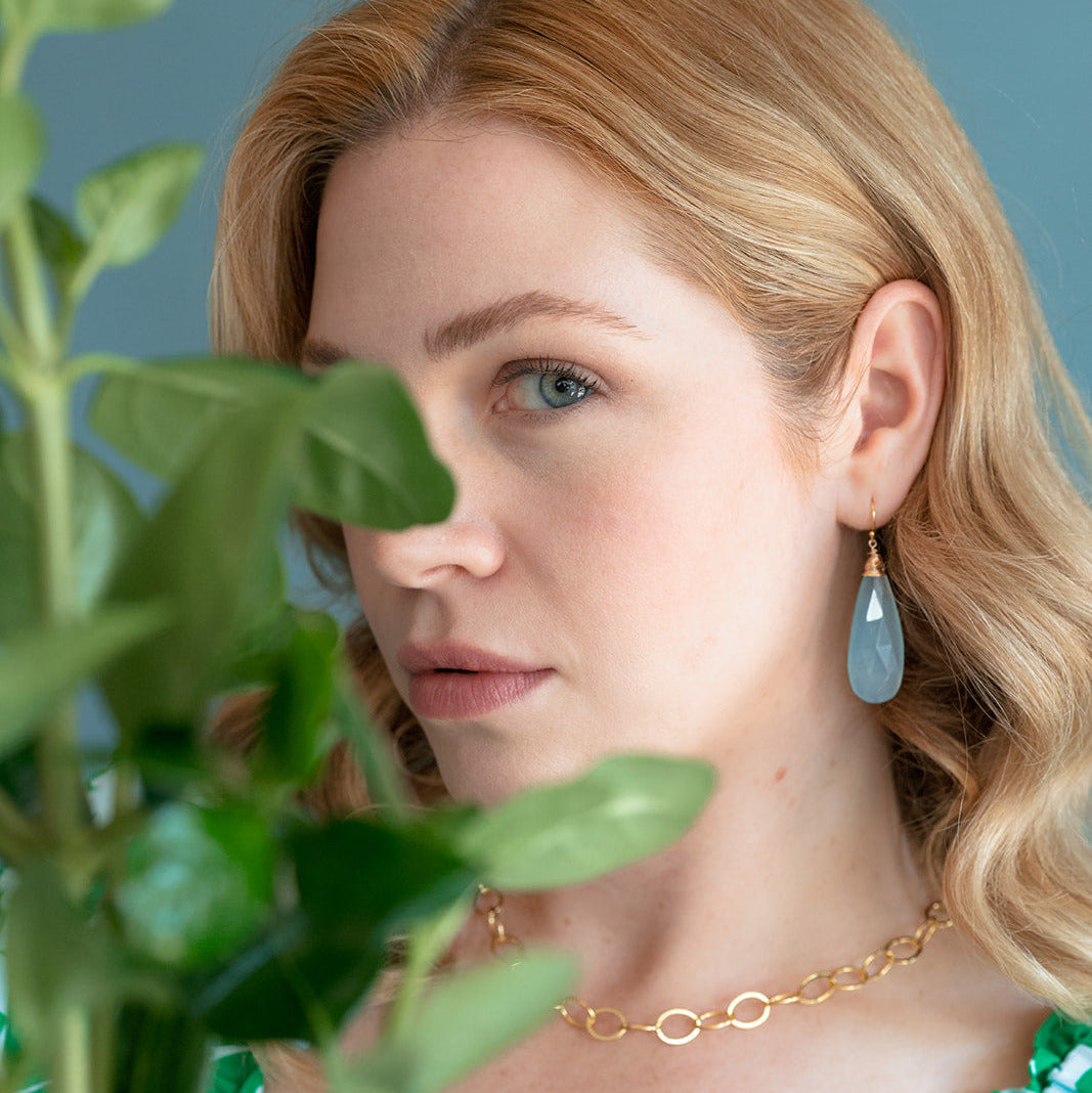 Woman wearing a gold necklace and green floral top with a plant in the foreground.