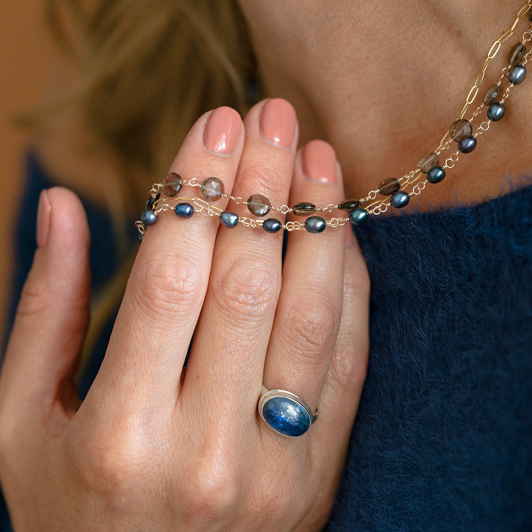 Woman holding a triple strand necklace draped over her back and wearing a blue sweater