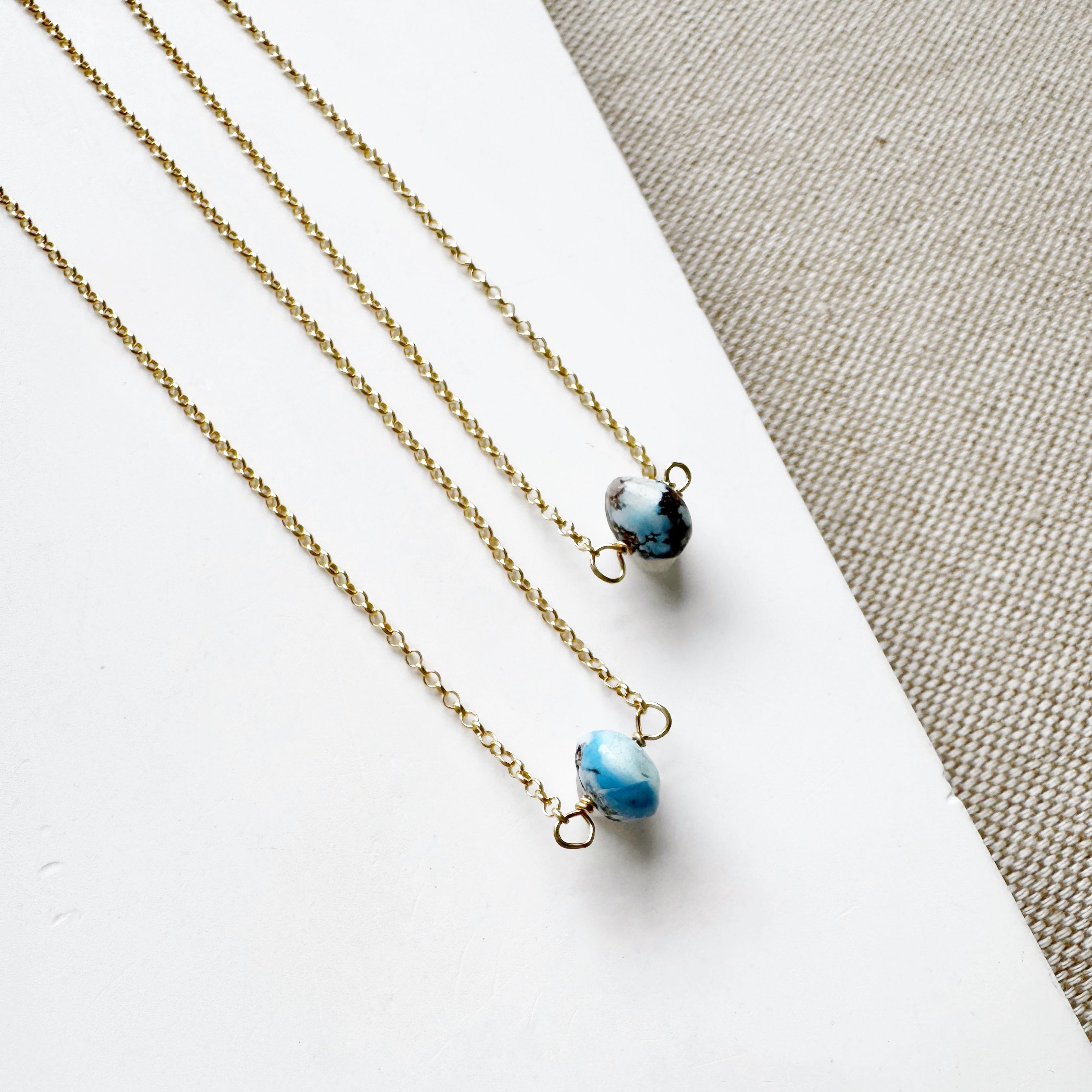 Two necklaces with a single small Golden Hill turquoise bead on gold chains on a white surface.