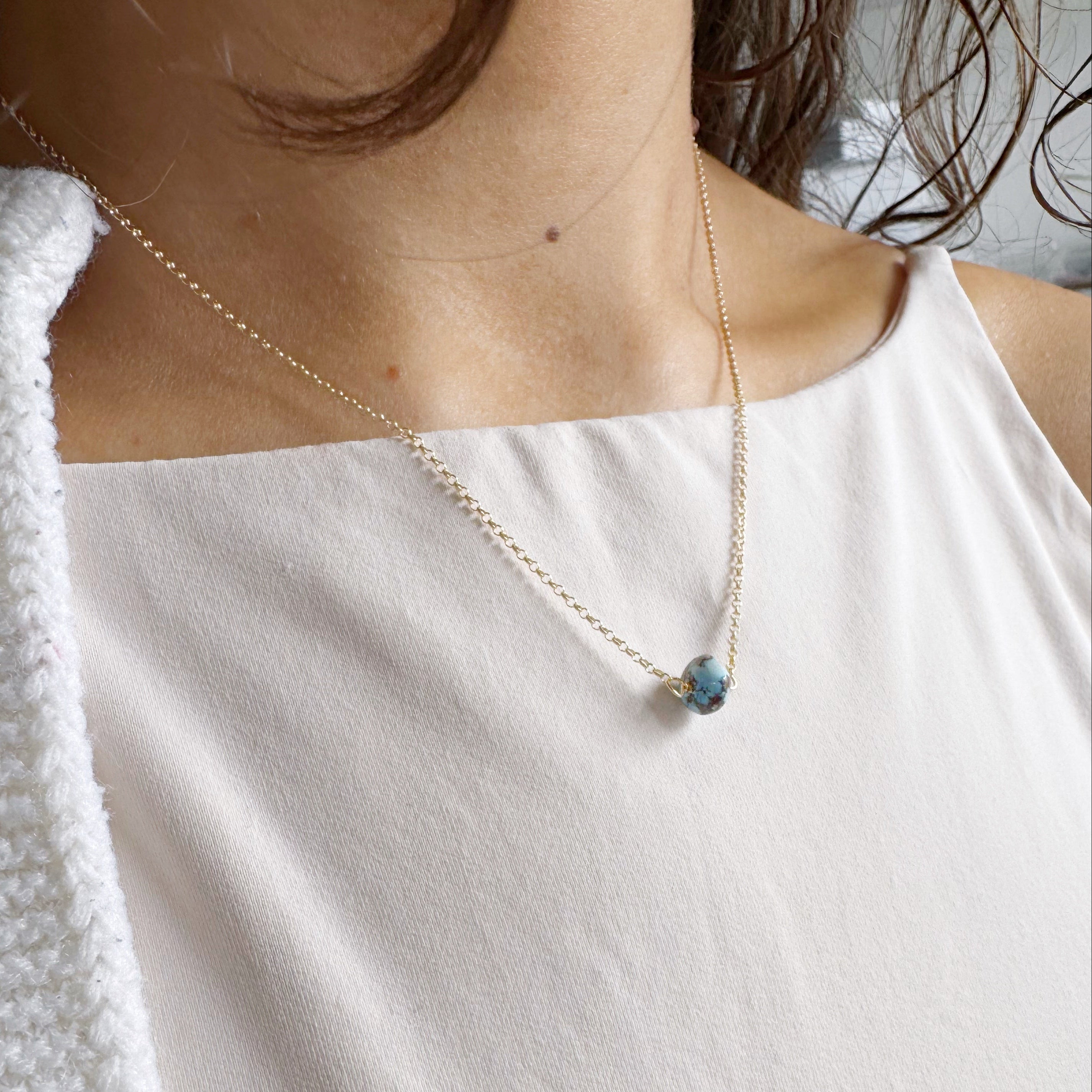 Necklace with a single small Golden Hill turquoise bead on gold chain worn by a person in a white top.