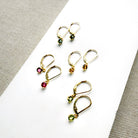 Four pairs of gold-toned leverback earrings with small round gemstones in green, yellow, and pink, arranged on a white surface with a textured gray fabric background.