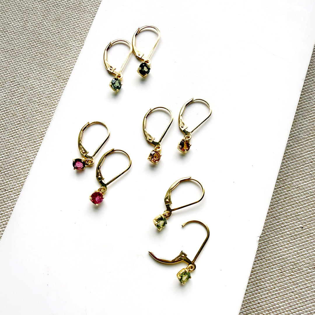 Four pairs of gold-toned drop earrings with small faceted gemstones in green, pink, orange, and yellow, arranged on a white rectangular surface with textured neutral fabric around the edges.