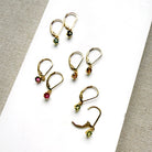 Four pairs of gold-toned drop earrings with small faceted gemstones in green, pink, orange, and yellow, arranged on a white rectangular surface with textured neutral fabric around the edges.