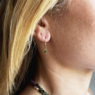 A close-up side profile of a person wearing a delicate gold huggie hoop earring with a small green faceted gemstone dangle, paired with a beaded necklace.