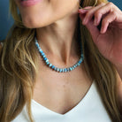 A close-up lifestyle shot of a woman wearing a chunky beaded necklace made of light blue and white marbled beads, paired with a white top and gold hoop earrings.