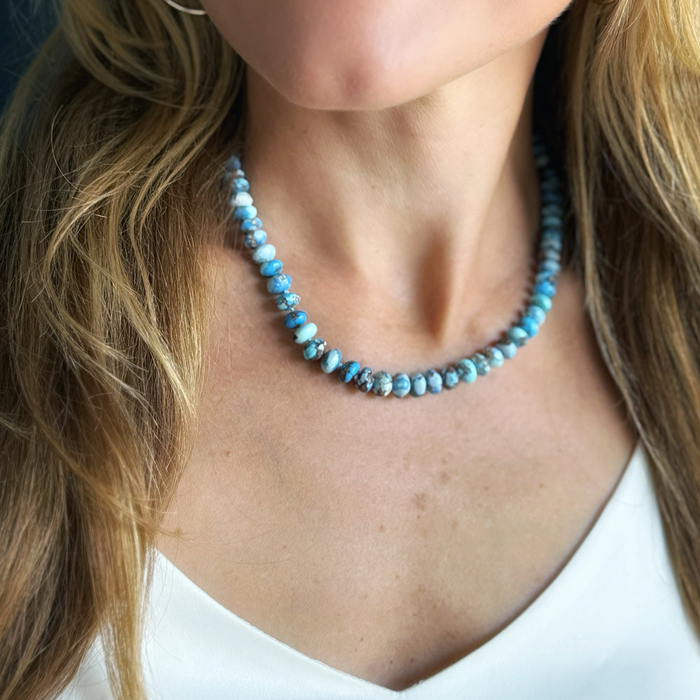 A close-up of a woman wearing a beaded necklace made of light blue and white marbled beads, paired with a white top.