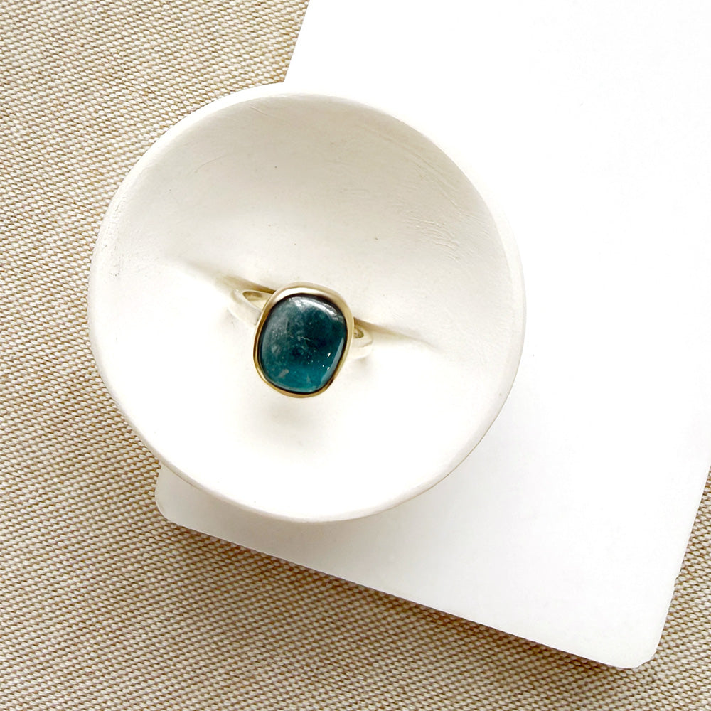 A silver ring with a small oval aquamarine cabochon gemstone, displayed on a white rectangular card over a beige woven fabric background.