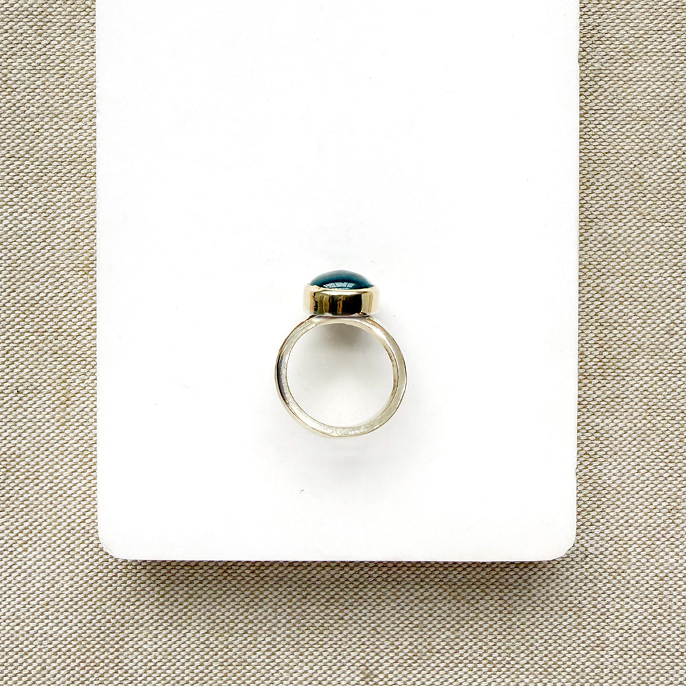 A silver ring with a small oval aquamarine cabochon gemstone, displayed on a white rectangular card over a beige woven fabric background.