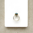 A silver ring with a small oval aquamarine cabochon gemstone, displayed on a white rectangular card over a beige woven fabric background.