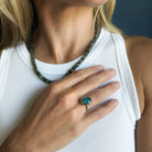 A close-up of a woman wearing a gold ring with an oval aquamarine cabochon stone, styled with a beaded necklace and a white ribbed tank top.