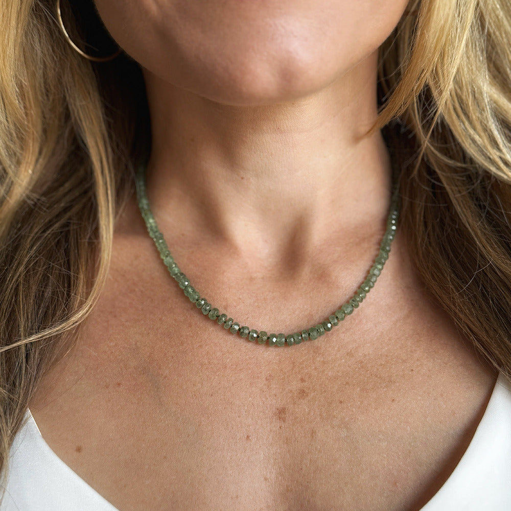 A close-up of a woman wearing a gemstone beaded necklace in green kyanite, paired with gold hoop earrings and a white top.