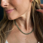 A close-up of a woman wearing a gemstone beaded necklace in green kyanite, paired with gold hoop earrings and a white top.