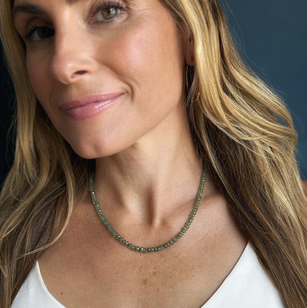A close-up of a woman wearing a gemstone beaded necklace in green kyanite, paired with gold hoop earrings and a white top.