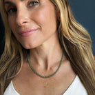 A close-up of a woman wearing a gemstone beaded necklace in green kyanite, paired with gold hoop earrings and a white top.
