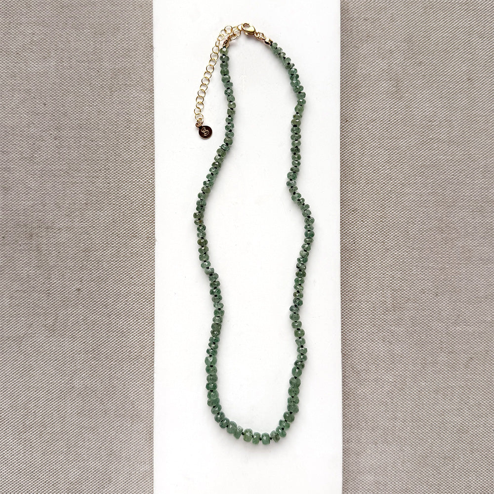 A beaded gemstone necklace made of green kyanite with a gold-tone clasp and extender chain, displayed on a white rectangular card against a textured gray fabric background.