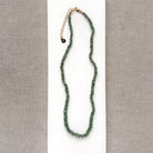 A beaded gemstone necklace made of green kyanite with a gold-tone clasp and extender chain, displayed on a white rectangular card against a textured gray fabric background.