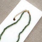 A beaded gemstone necklace made of green kyanite with a gold-tone clasp and extender chain, displayed on a white rectangular card against a textured gray fabric background.