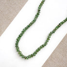 A beaded gemstone necklace made of green kyanite with a gold-tone clasp and extender chain, displayed on a white rectangular card against a textured gray fabric background.