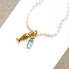 A white pearl beaded necklace featuring a gold-toned fish charm and a small light blue rectangular stone pendant, displayed on a white card over a textured beige fabric background.