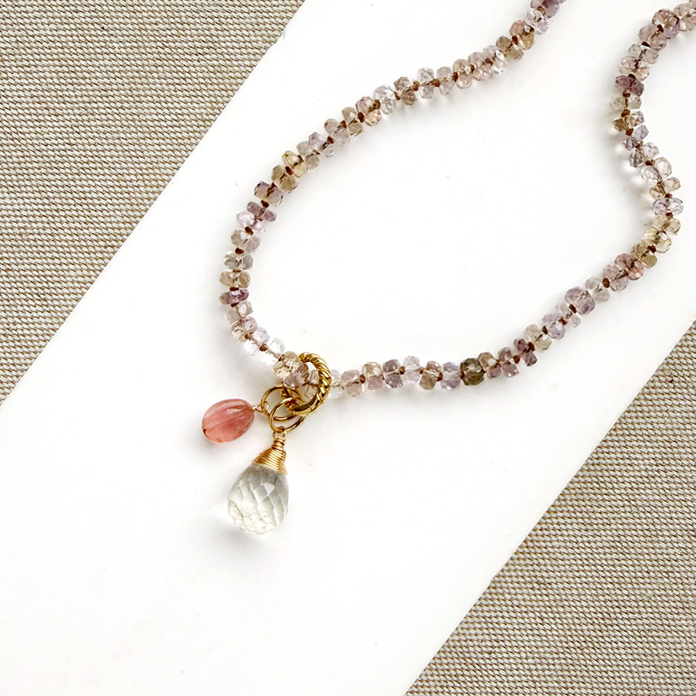 A beaded necklace with small faceted beads in pink, brown, and clear tones, featuring a gold-tone clasp and two dangling charms: a pink teardrop bead and a larger clear faceted bead, displayed on a white card over a beige woven fabric background.