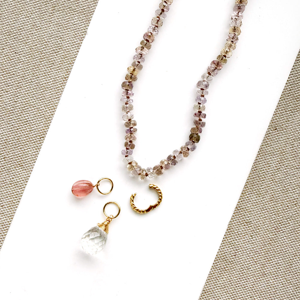 A beaded necklace with faceted crystal beads in soft pink, purple, and brown tones, featuring a gold-tone pendant with a pink teardrop bead, a clear crystal teardrop, and a small gold hoop, displayed on a white card over a beige woven fabric background.