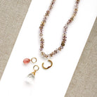A beaded necklace with faceted crystal beads in soft pink, purple, and brown tones, featuring a gold-tone pendant with a pink teardrop bead, a clear crystal teardrop, and a small gold hoop, displayed on a white card over a beige woven fabric background.