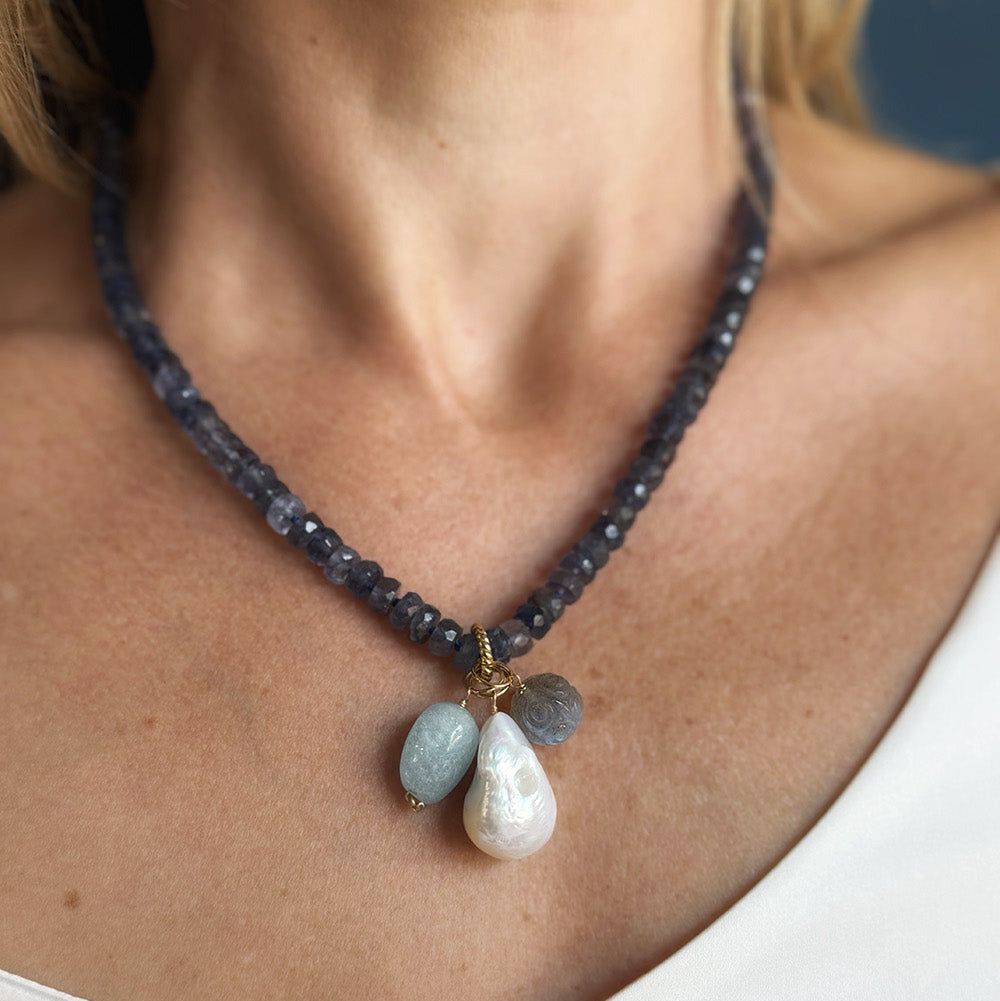 A close-up of a person wearing a beaded necklace with dark faceted beads and three teardrop-shaped stone pendants in pale blue, gray, and white, styled with a gold accent.
