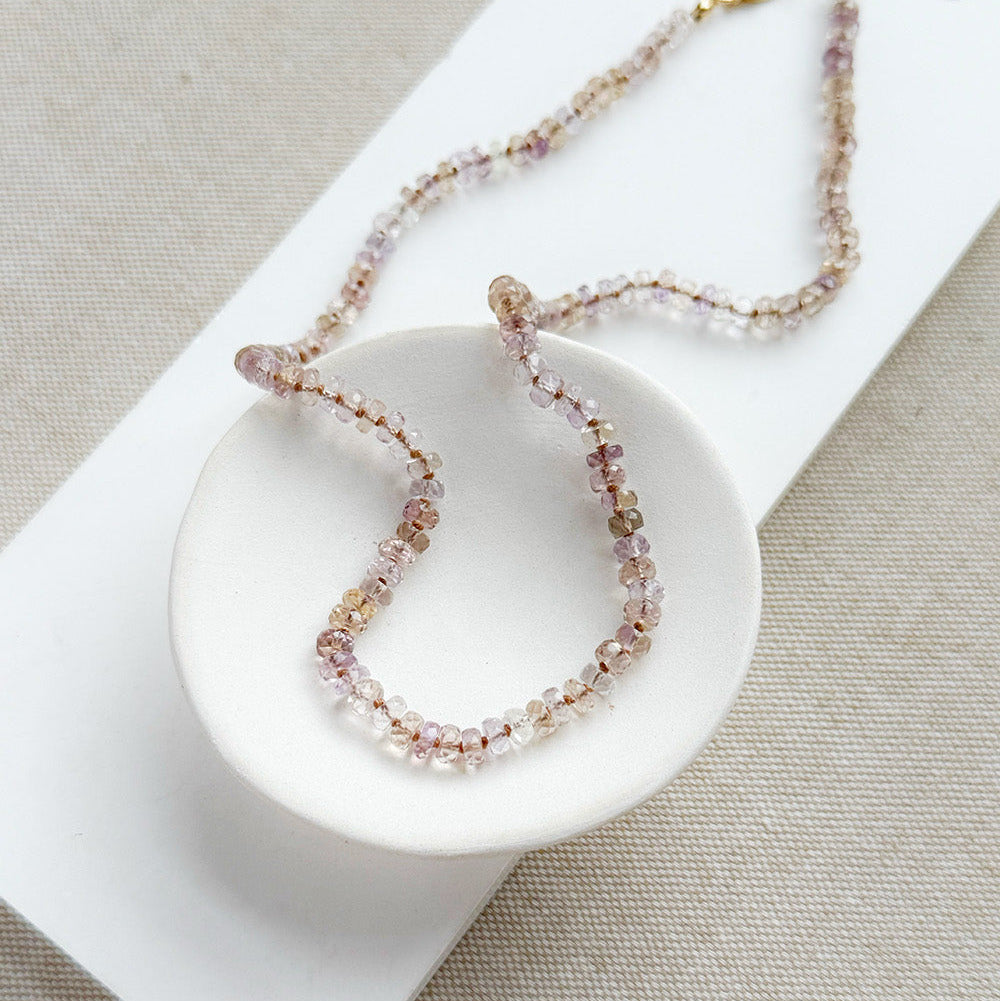 A delicate beaded necklace with small pink, peach, and clear faceted beads, accented by a gold clasp, displayed on a small white dish over a beige textured fabric background.