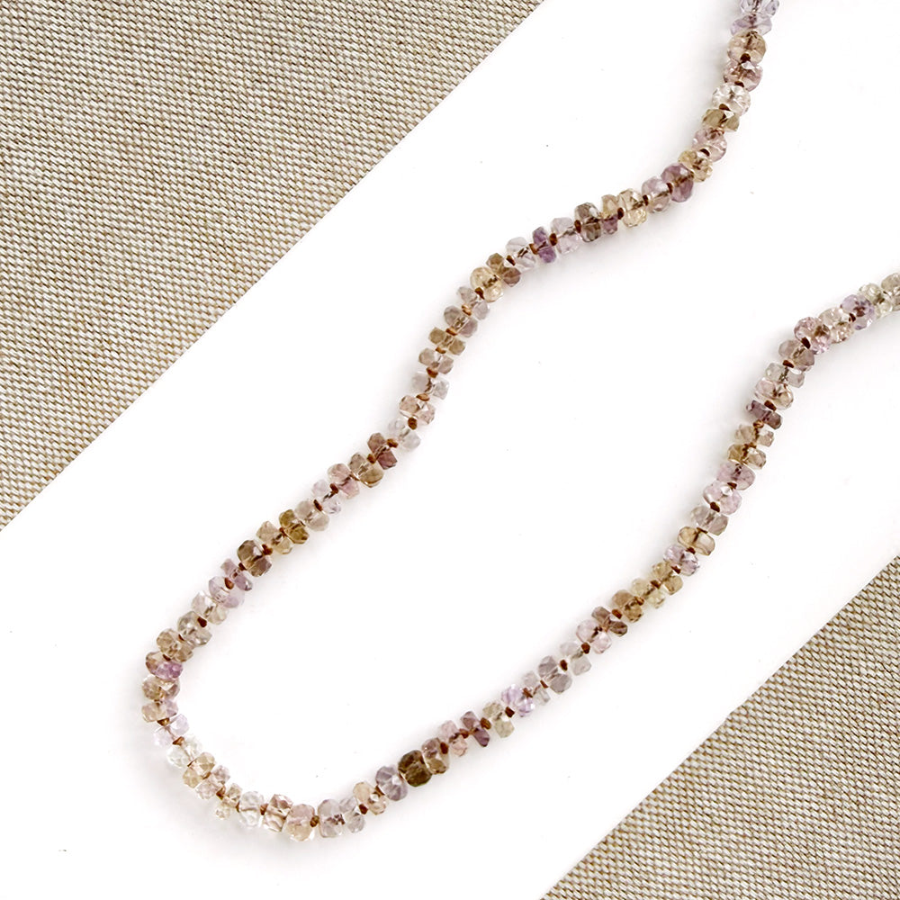 A strand of small faceted gemstone beads in a multicolor mix of pink, purple, brown, and clear tones, arranged on a white surface with a beige woven fabric background.