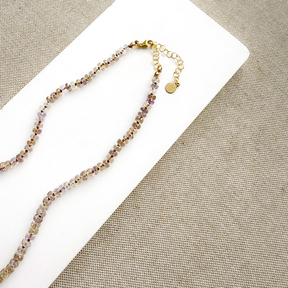 A delicate beaded necklace with faceted brown and lavender beads on a gold chain, displayed on a white rectangular card over a beige woven fabric background.
