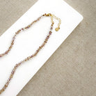 A delicate beaded necklace with faceted brown and lavender beads on a gold chain, displayed on a white rectangular card over a beige woven fabric background.