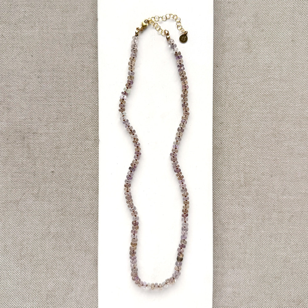 A delicate beaded necklace with small lavender, purple, and brown beads, featuring a gold-tone clasp and extender chain, displayed on a white card against a textured gray background.