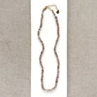A delicate beaded necklace with small lavender, purple, and brown beads, featuring a gold-tone clasp and extender chain, displayed on a white card against a textured gray background.