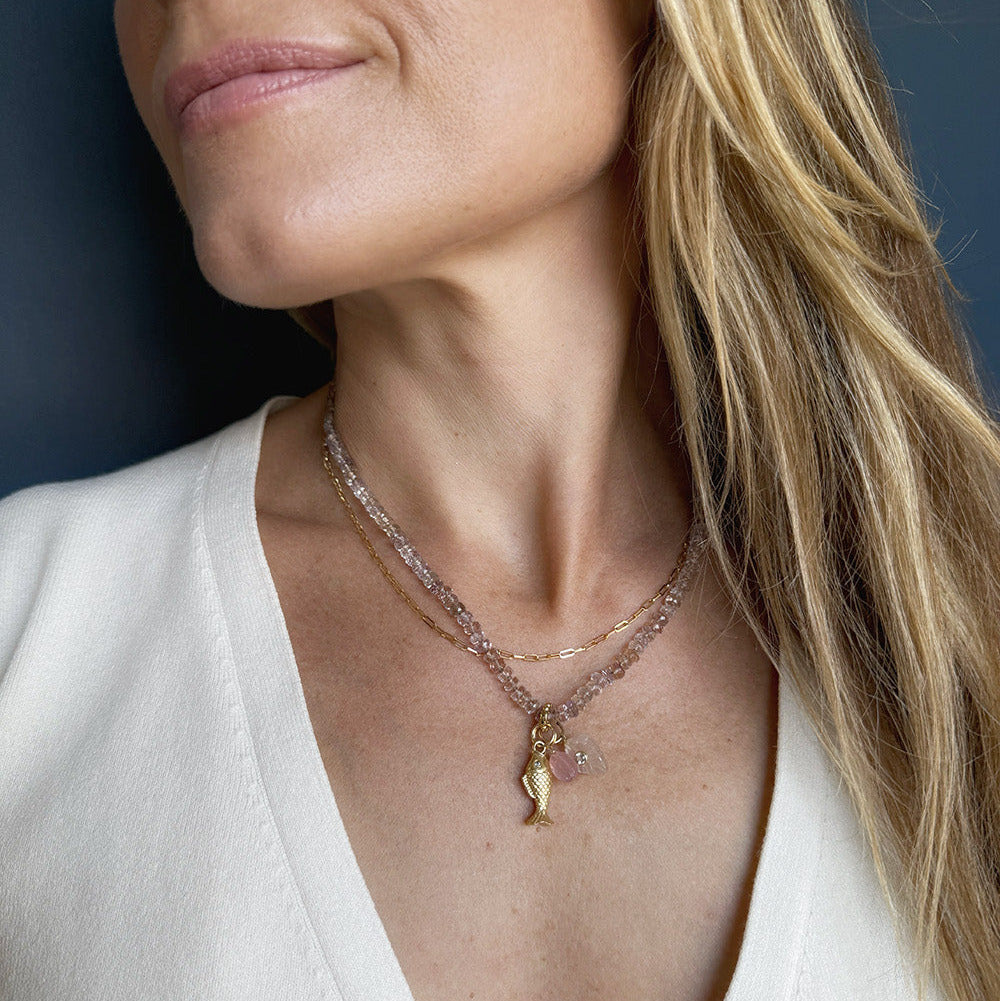 A close-up of a woman wearing a delicate layered necklace with a small gold pendant and pink gemstone charms, paired with a white V-neck top against a dark blue background.