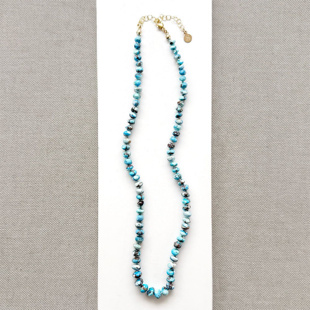 A knotted Golden Hills turquoise necklace, draped over a white plate on a neutral textured surface.
