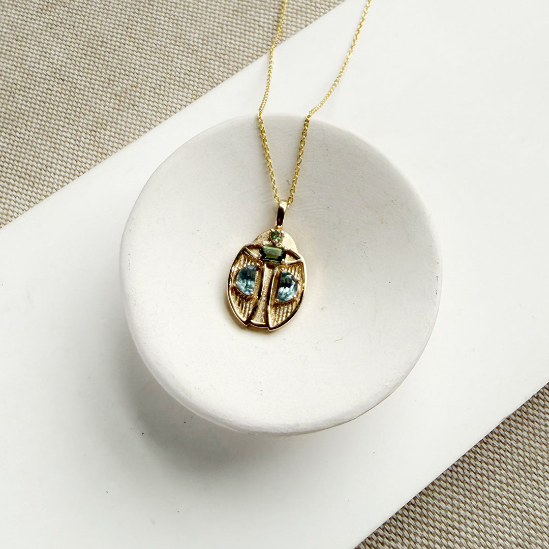 Gold beetle pendant necklace with light blue gemstones on a white ceramic dish, set against a textured beige and white background.
