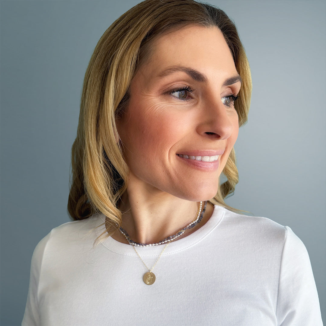 A blonde woman wearing a gold pendant necklace with an engraved flower on a white shirt with a light blue background