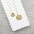 Two gold necklaces with round pendants, one small and one large, on a white stand against a gray background