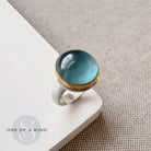Ring with a blue aquamarine stone on a beige surface, featuring 'One of a Kind' branding.