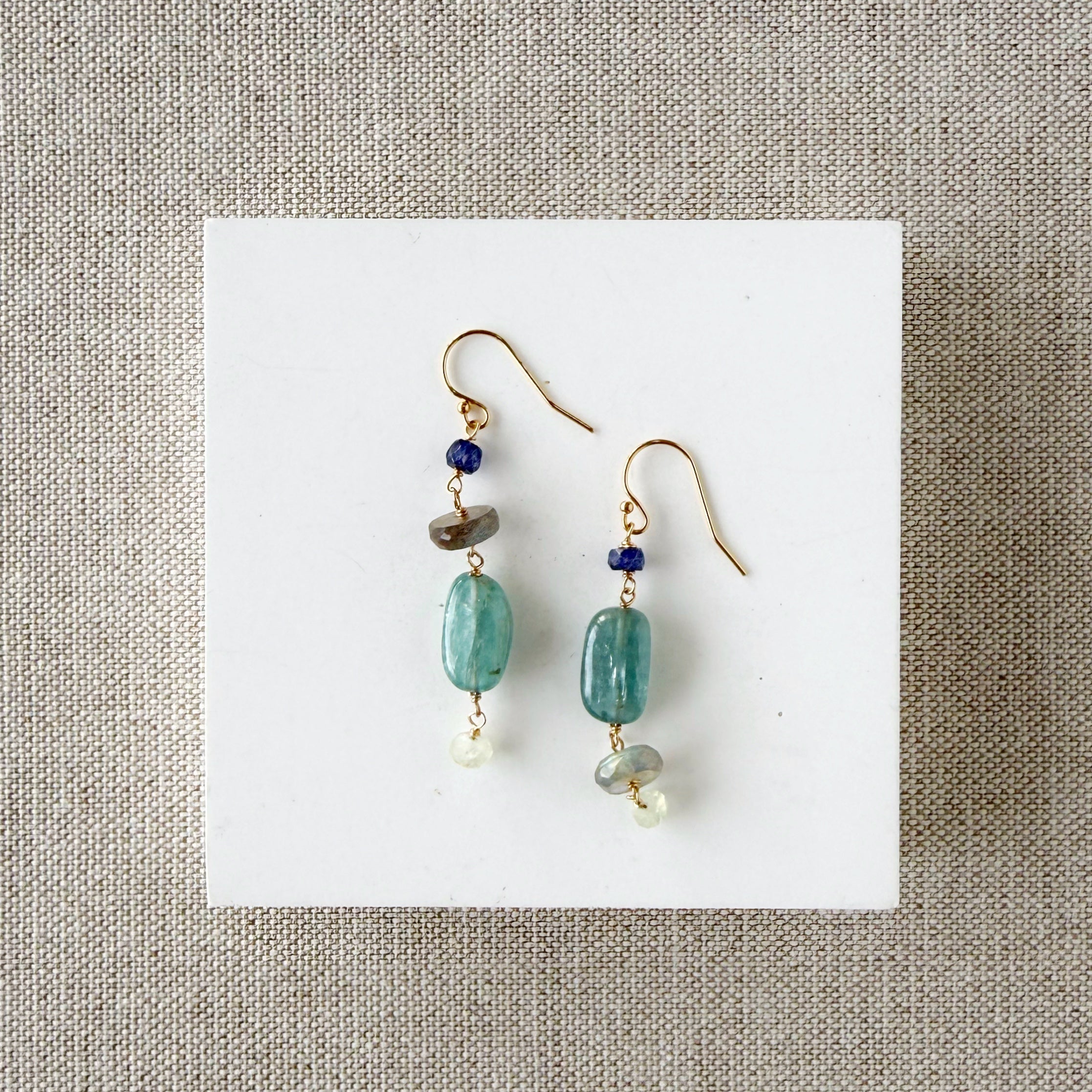 Pair of earrings with blue and teal gemstones on a white card against a beige background